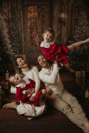A family wearing white and red outfits plays and laughs together during a holiday photo session against a rustic backdrop.