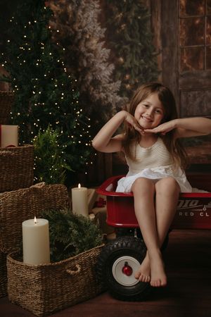 A child sits on a red bench near a Christmas tree with glowing lights and rustic holiday decor in the background.