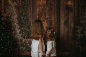 Romantic winter wedding couple embraces in front of rustic wooden wall with twinkling lights and greenery.