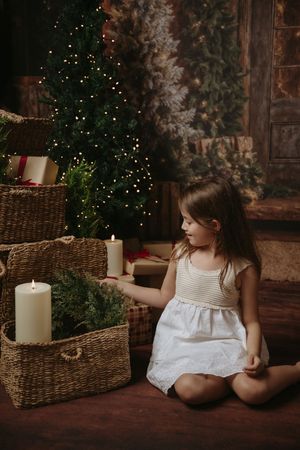 A child in a white dress sits near a Christmas tree with glowing lights and wicker baskets with candles.