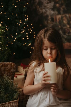 A child in a white dress sits in a cozy setting with twinkling lights and a lit candle creating a magical holiday atmosphere.