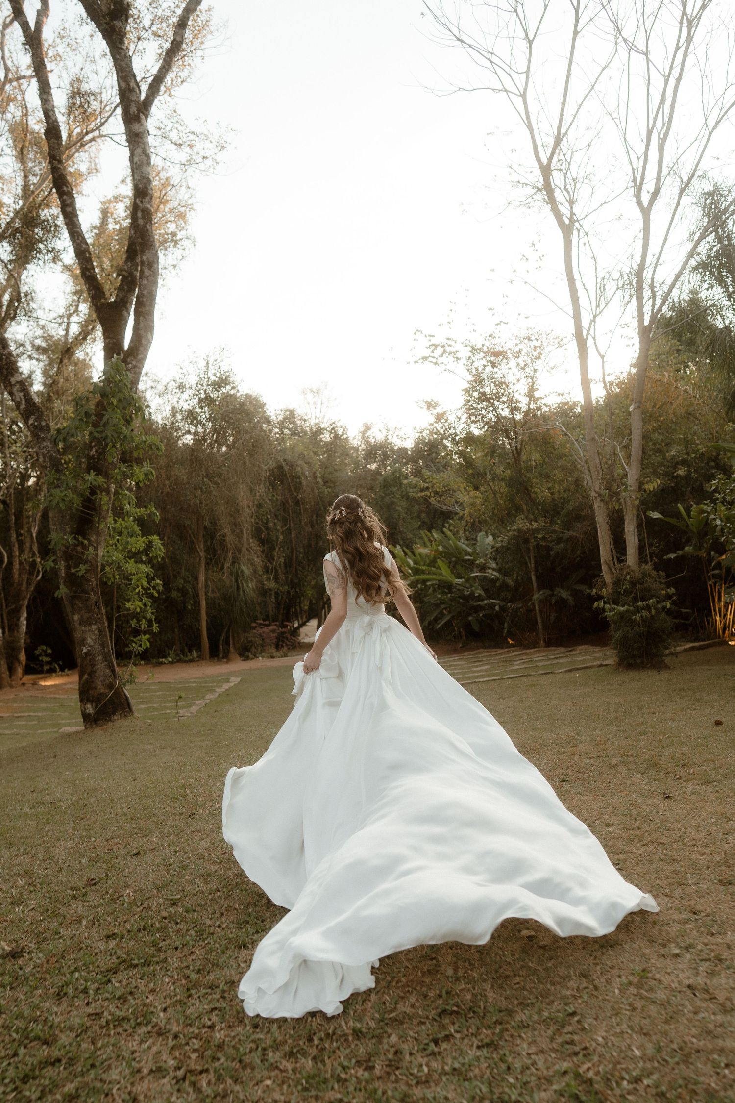 A flowing white wedding dress billows dramatically in the wind as someone spins in a grassy field at sunset.