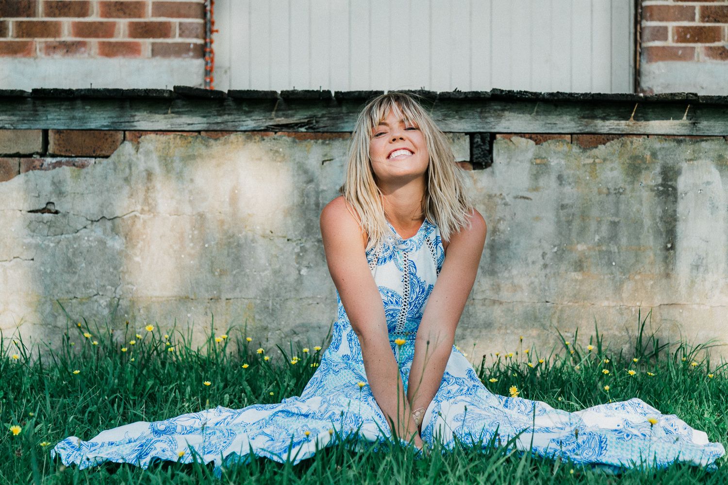 Woman in blue floral dress lying on grass near brick wall outdoors.