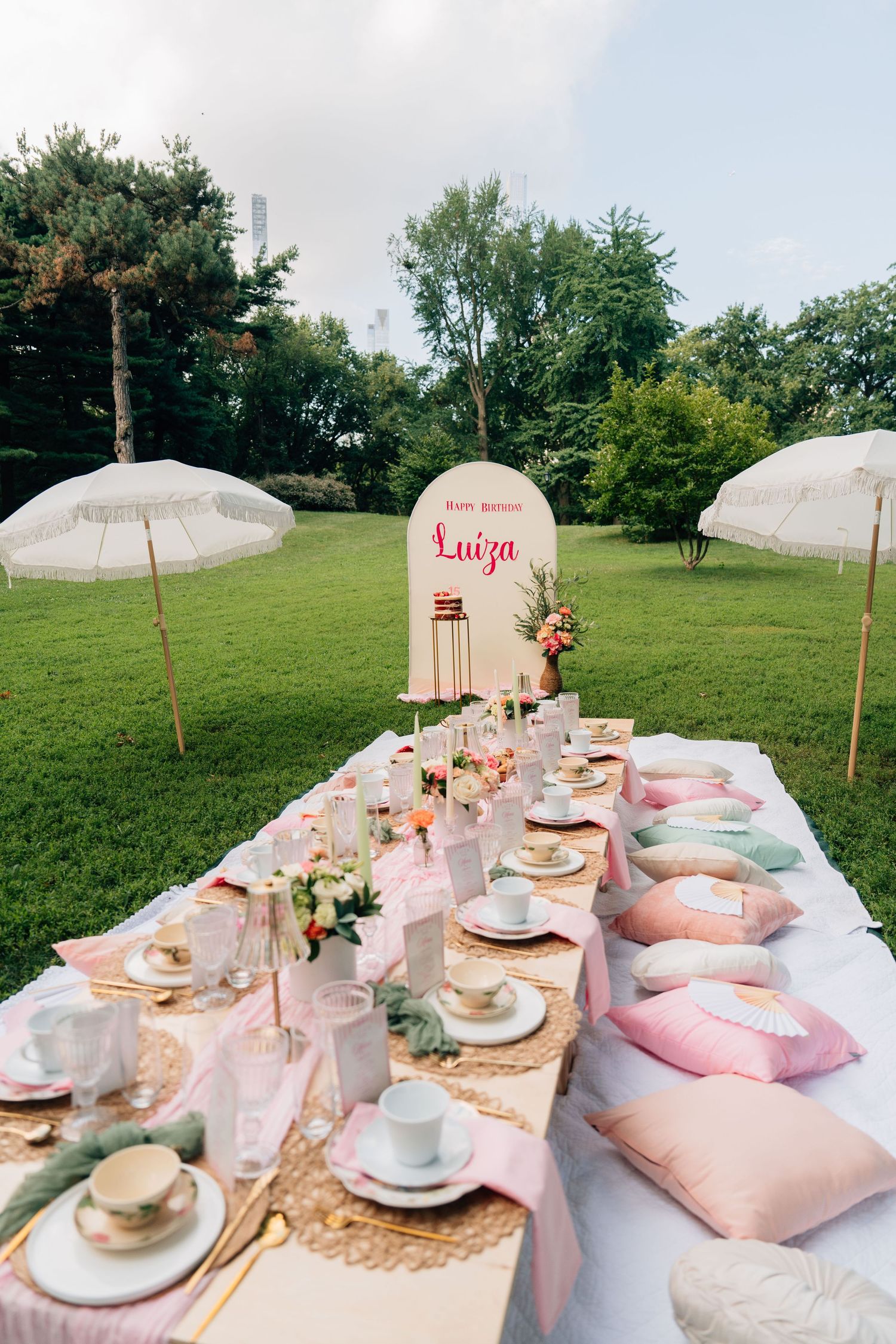 Elegant outdoor picnic setup with white umbrellas, pink tablecloths, and vintage teacups arranged on grassy hillside.