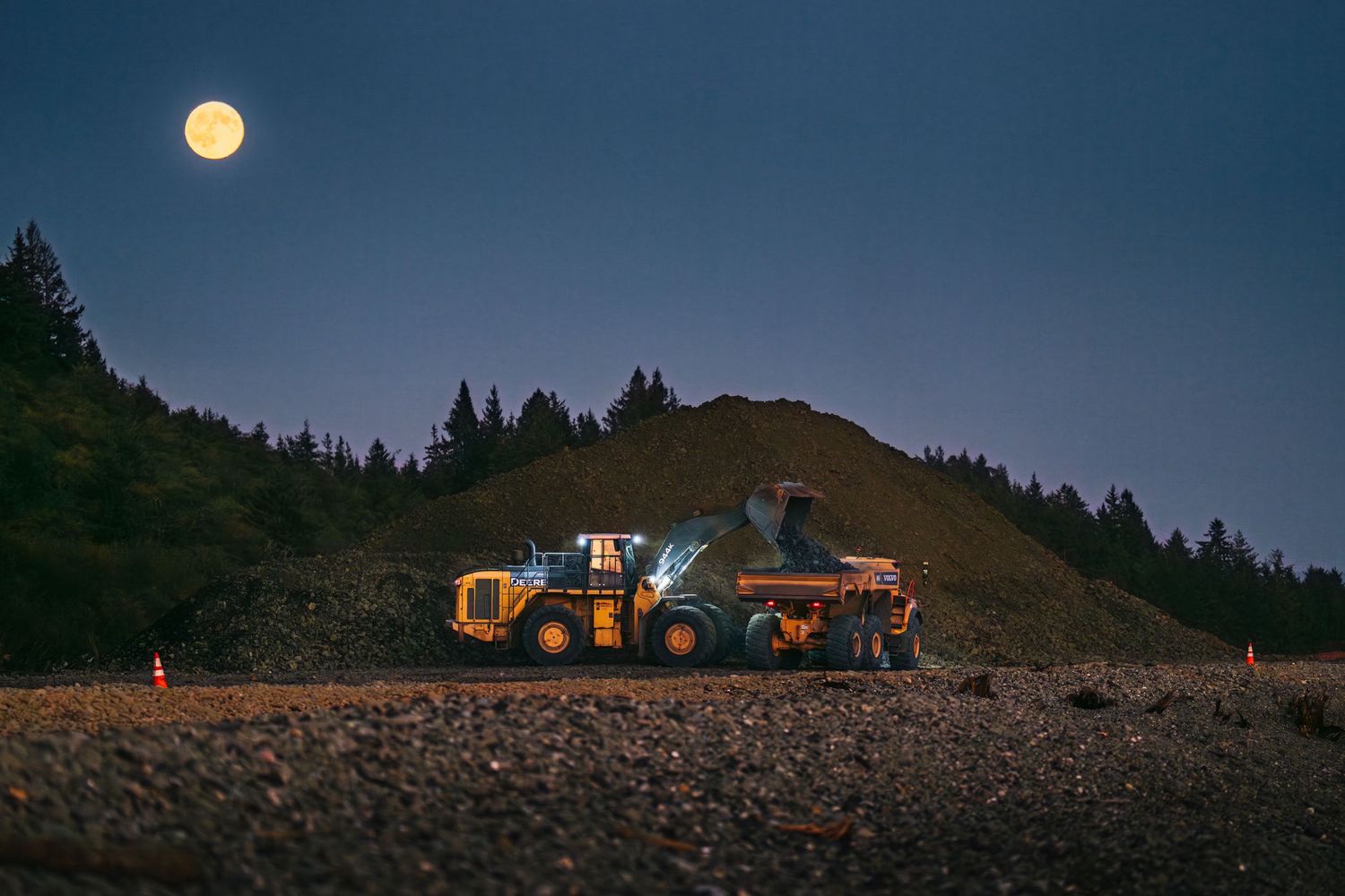 Yellow excavators working on industrial site against sunset sky with industrial buildings in background.