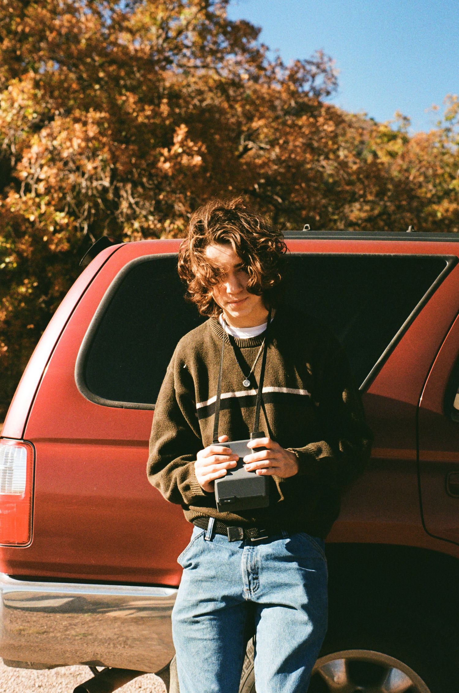 A person in a striped sweater and jeans stands next to a red vehicle on an autumn day with golden leaves in the background.
