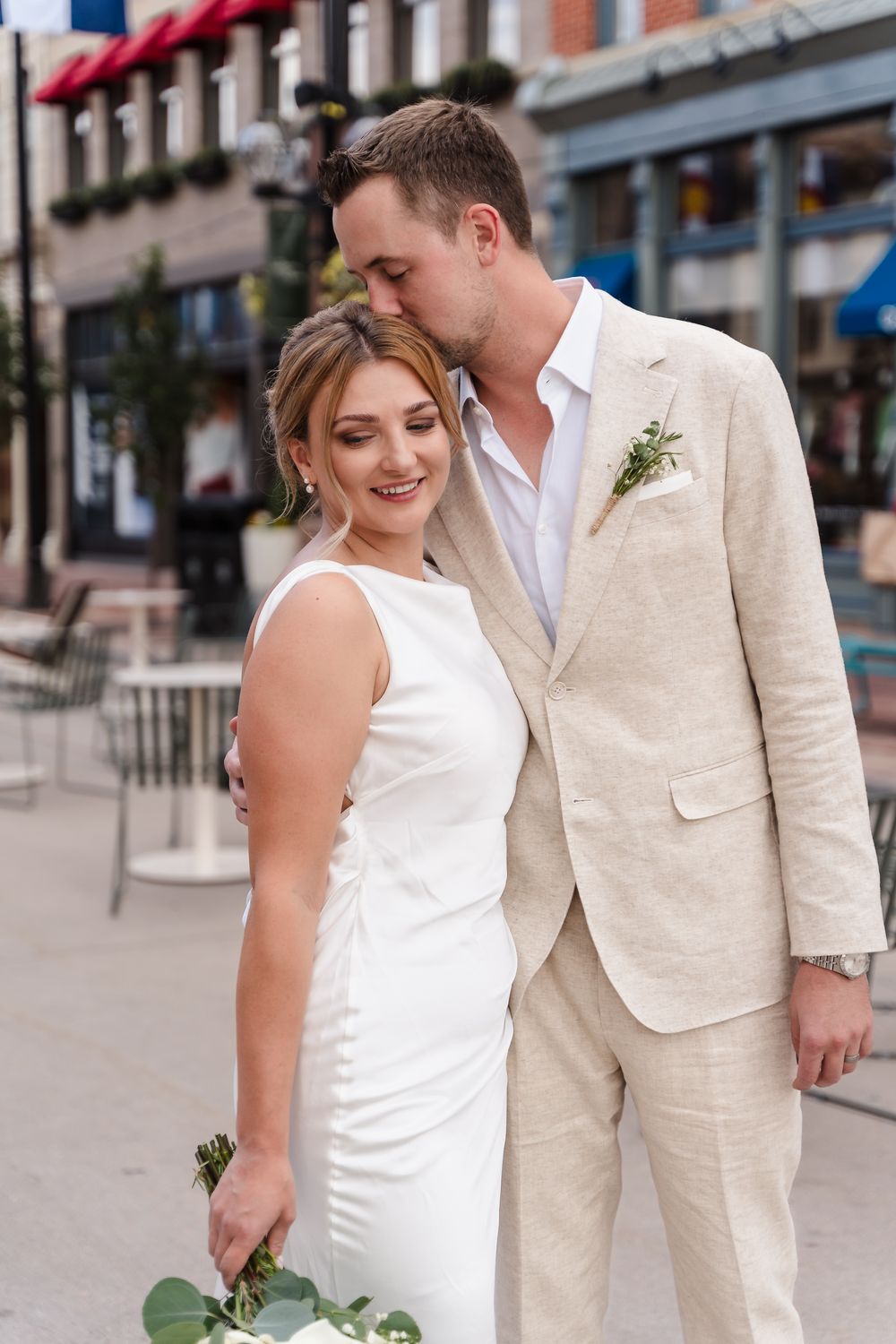 A couple in wedding attire shares an intimate moment on a city street with brick buildings in the background.