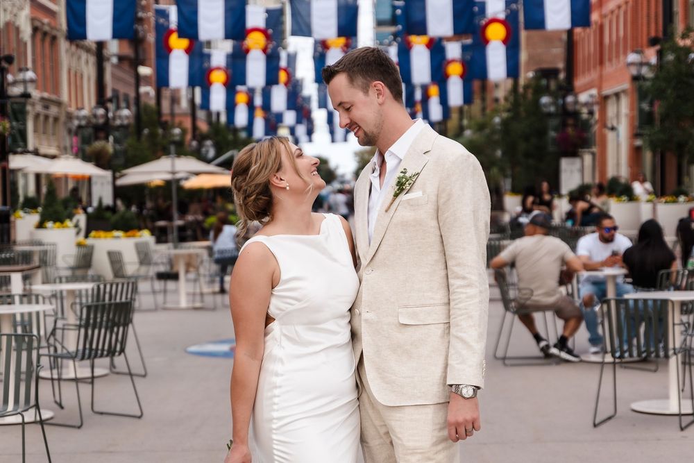 A couple in light colored attire shares a romantic moment under orange and blue decorative flags at an outdoor venue.