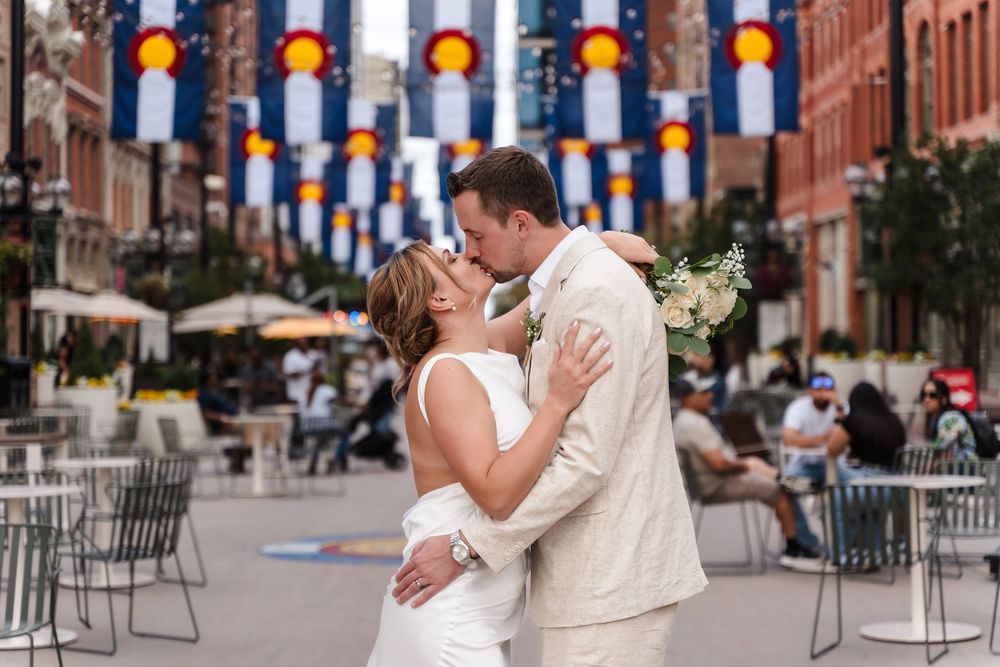 A couple shares a romantic kiss during their wedding celebration in an urban setting with string lights overhead.