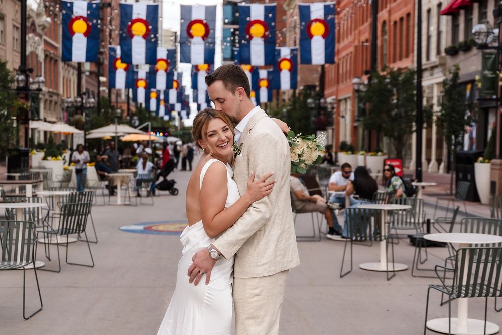 Colorful flags and banners hang over a brick-lined street with outdoor dining tables in a city neighborhood.