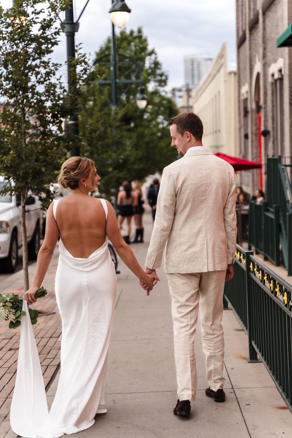 A couple in wedding attire walks hand in hand down a city sidewalk on their special day.