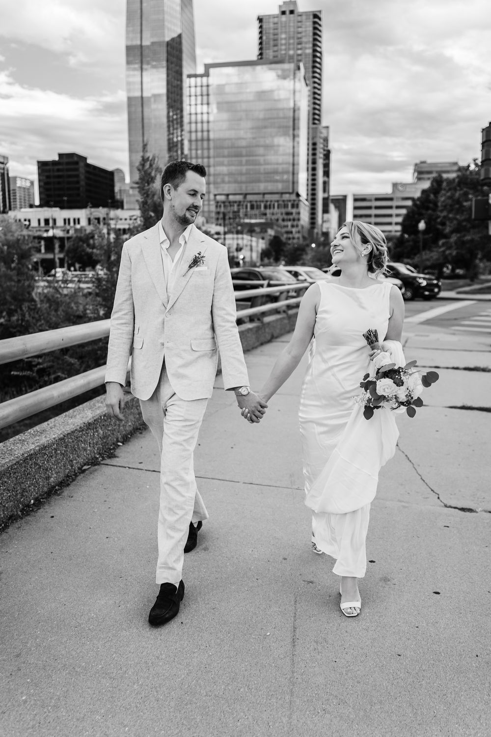 A black and white wedding photo of a couple in white attire walking together on a city bridge with skyscrapers in background.