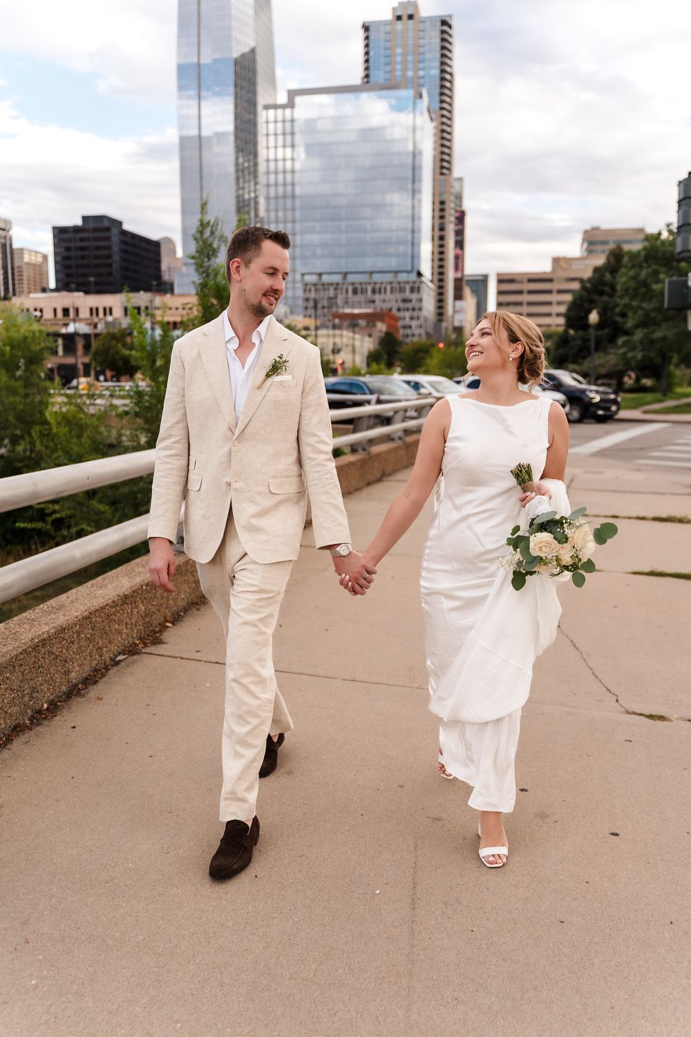 A couple in matching cream-colored wedding attire walks together on a paved path with city buildings in the background.