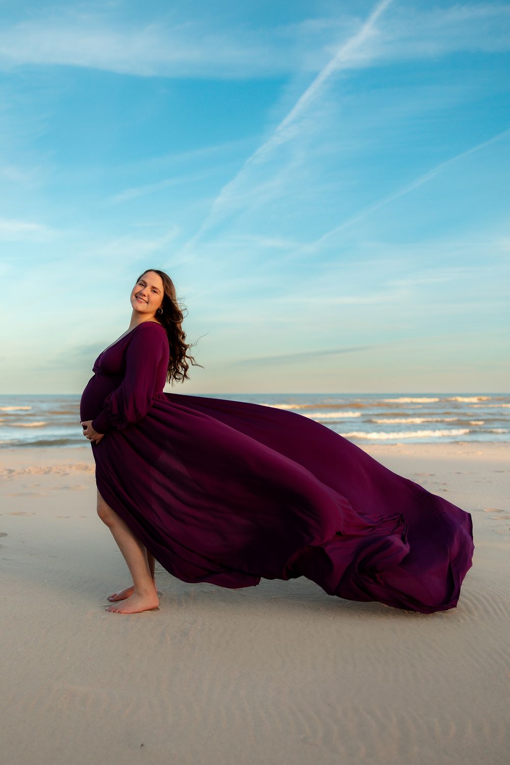 A flowing burgundy dress billows in the wind as someone poses on a sunny beach with waves and blue sky in the background.