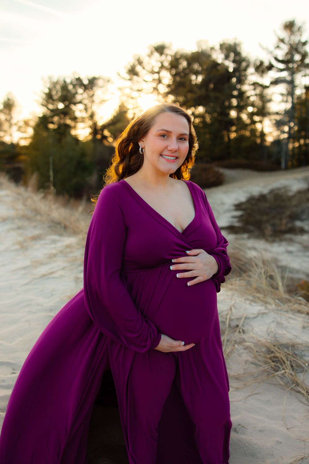A person in a flowing purple maternity dress poses at sunset in a natural outdoor setting with tall grass.