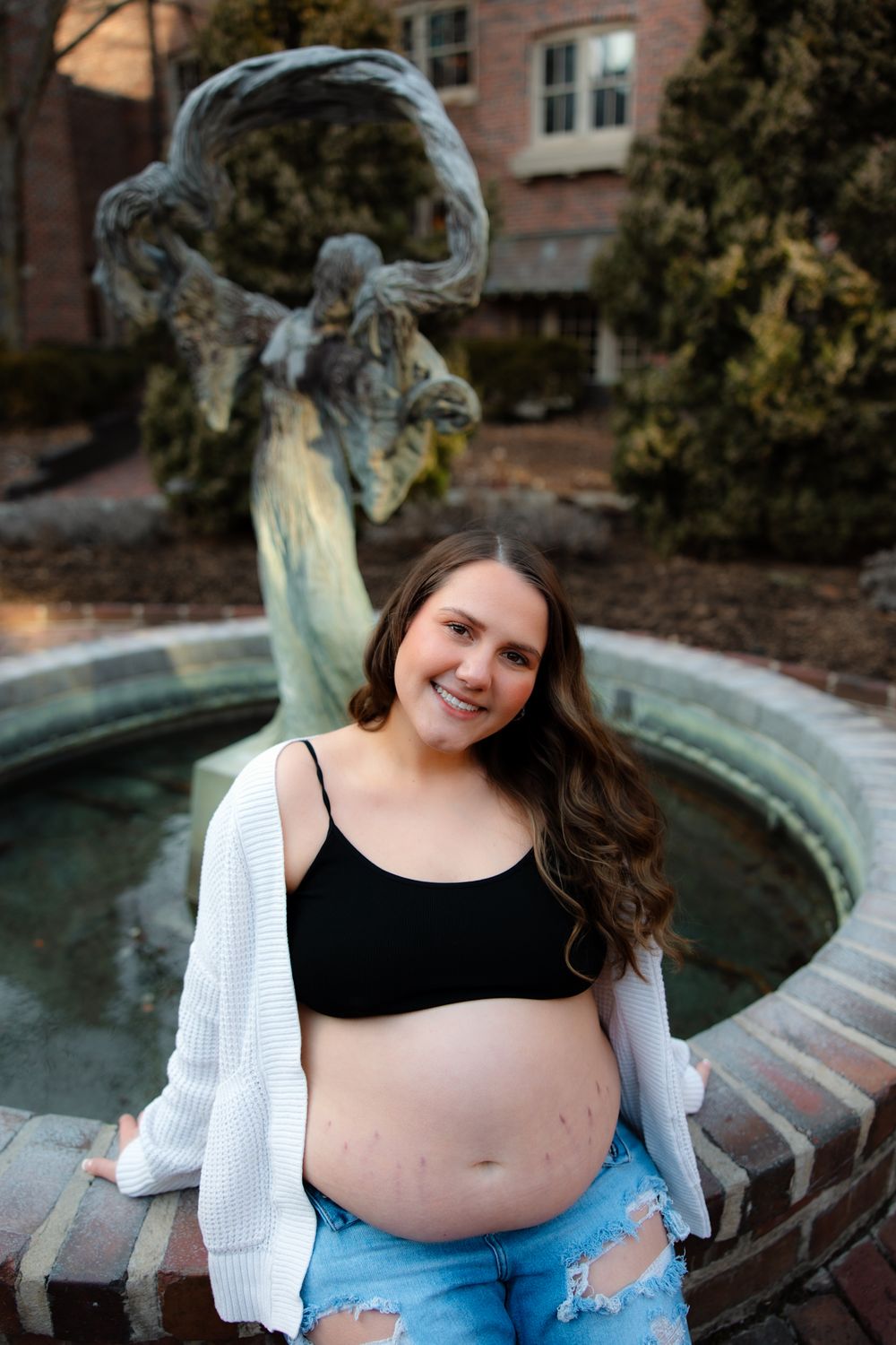 A pregnant woman in a blue dress sits by a stone fountain in an outdoor setting.