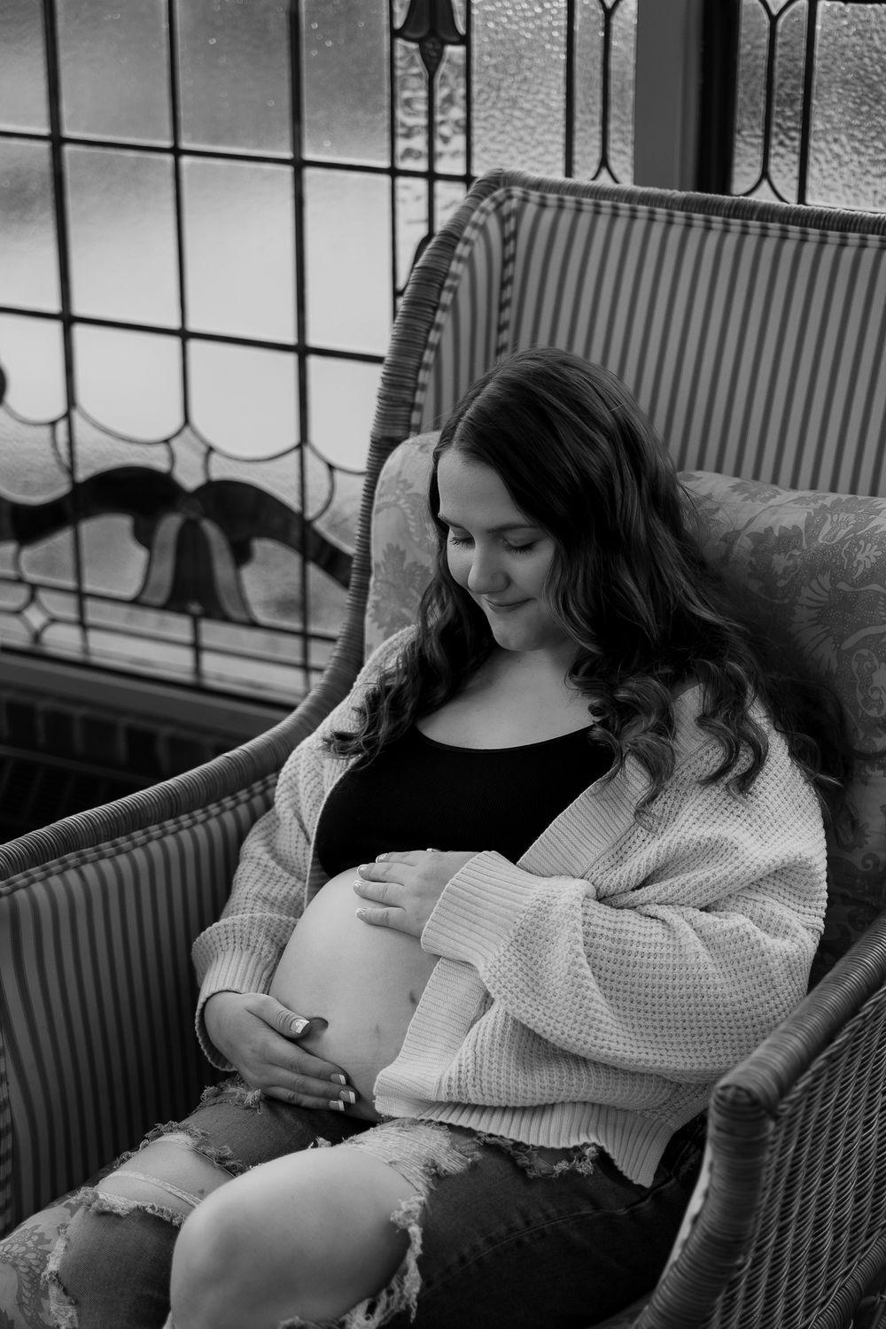 A black and white maternity photo showing an expectant mother relaxing in a wicker chair.