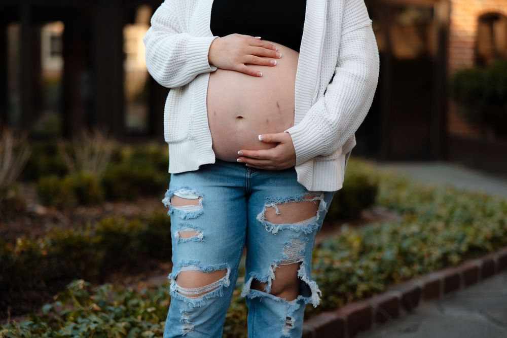 A close-up view of a pregnant belly with hands resting on it, wearing ripped jeans and a white sweater.