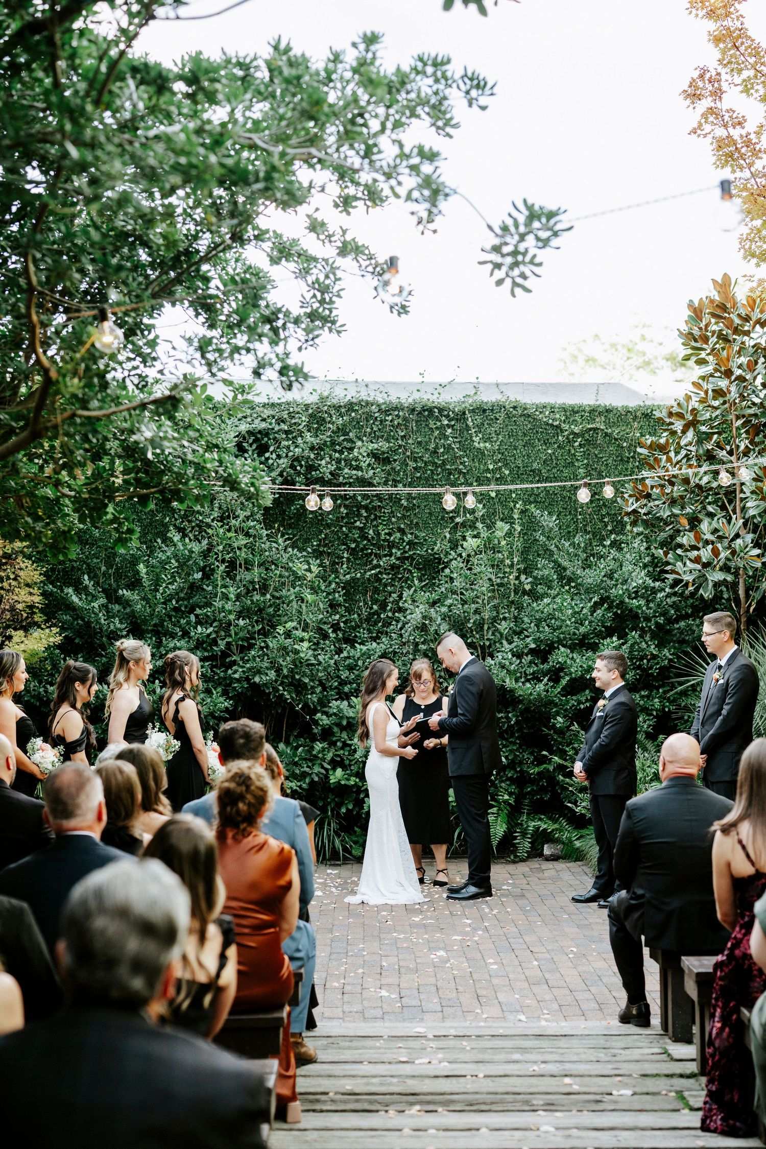 Guests gather outdoors for a wedding ceremony on a brick patio surrounded by lush greenery and trees.