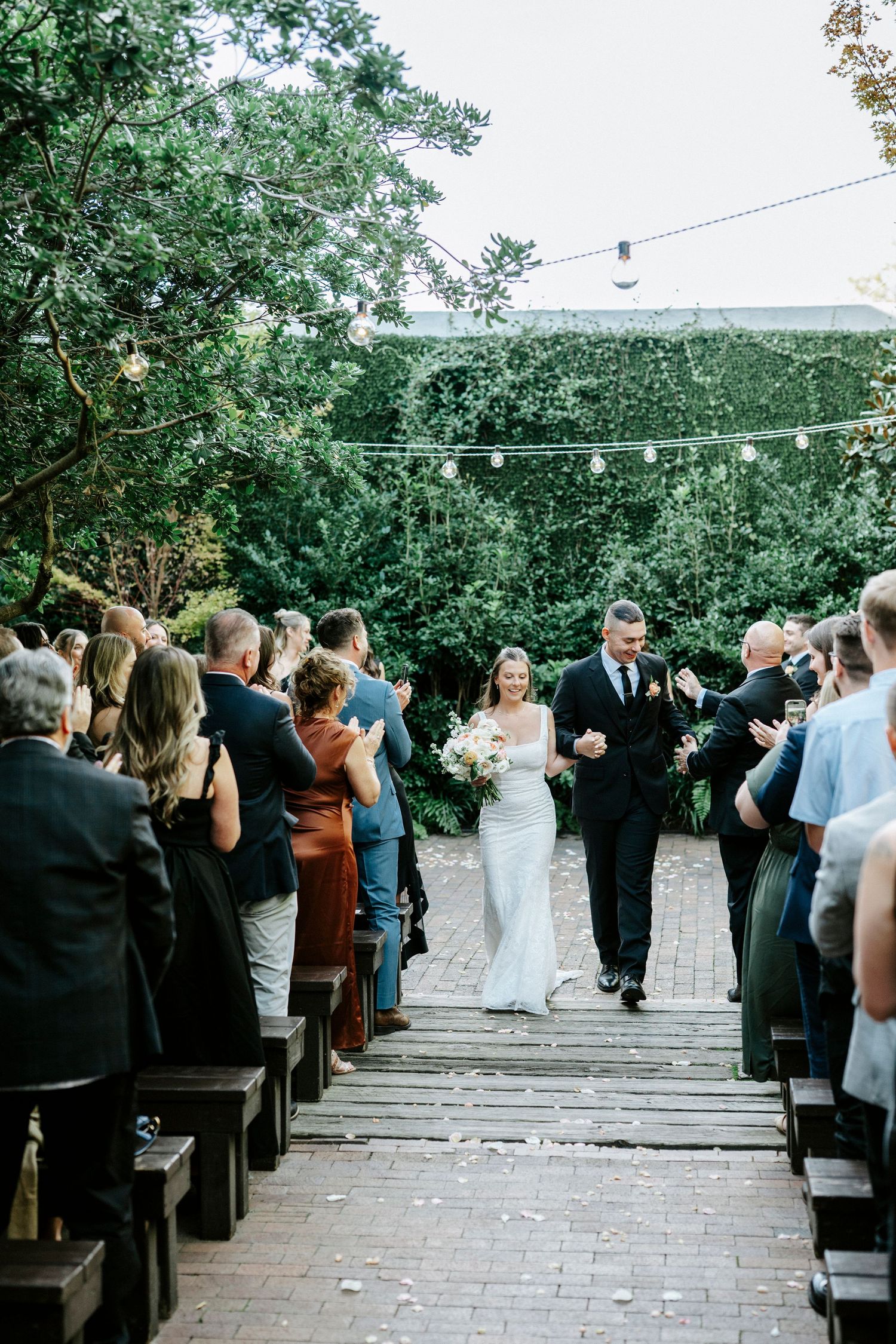 Outdoor wedding ceremony setup with rows of wooden benches and string lights in a garden setting.