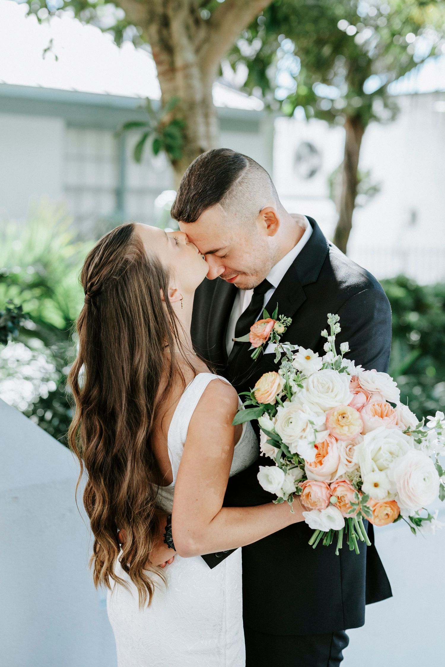 Romantic wedding couple shares intimate moment with peach and white floral bouquet against white backdrop.