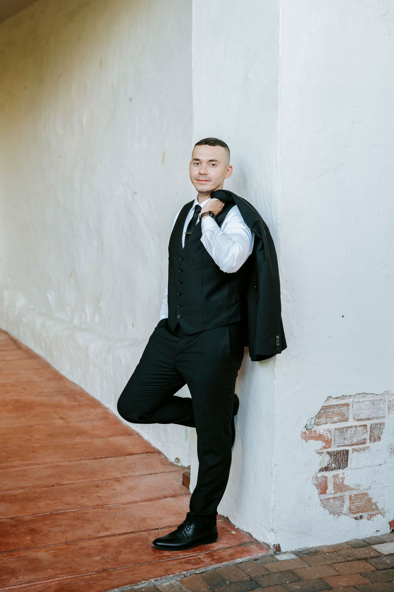 Stylish groom in black suit poses against white wall during wedding photoshoot.