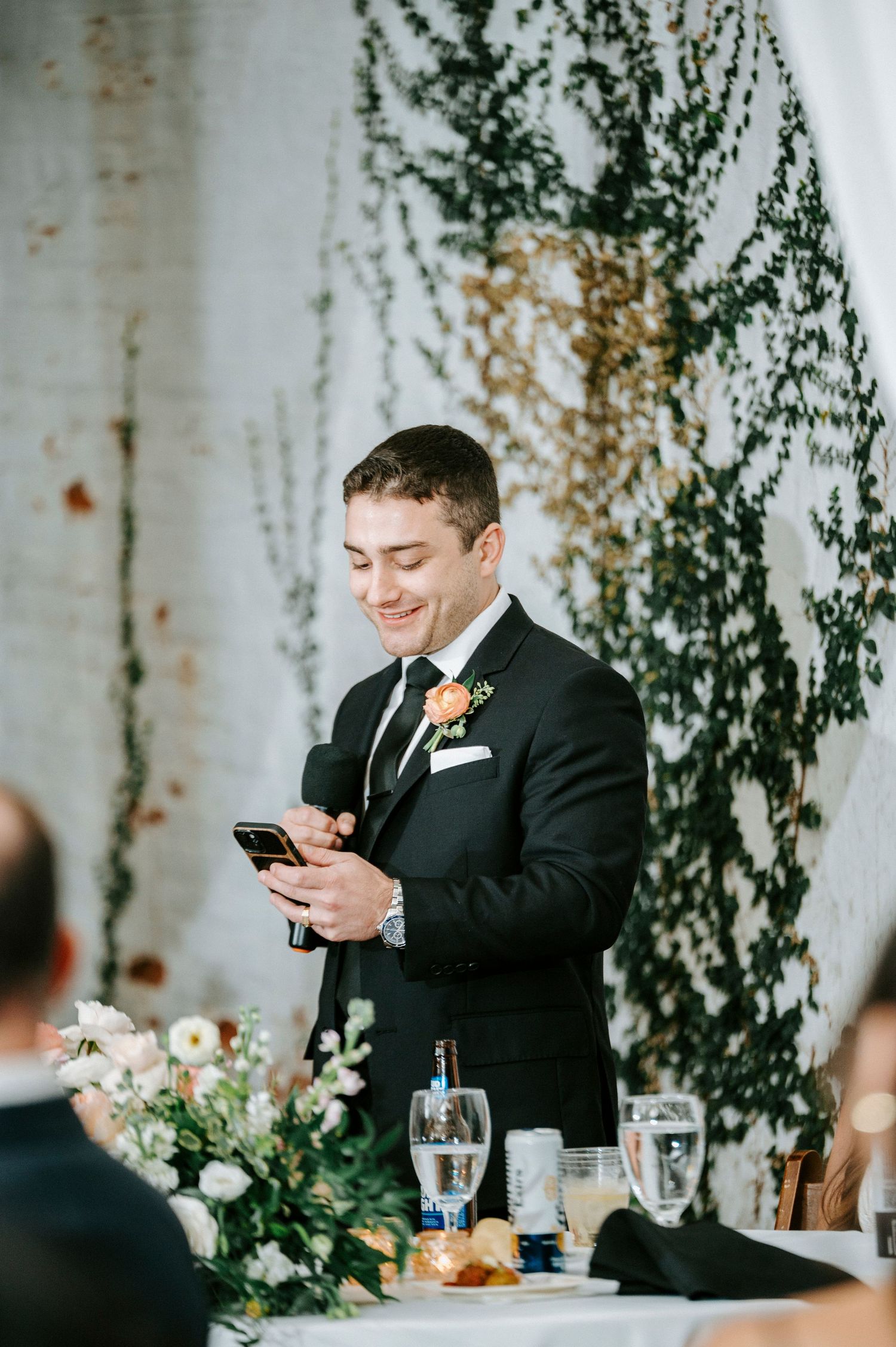 A person in a black suit smiles while looking at their phone during a formal event with greenery decor.