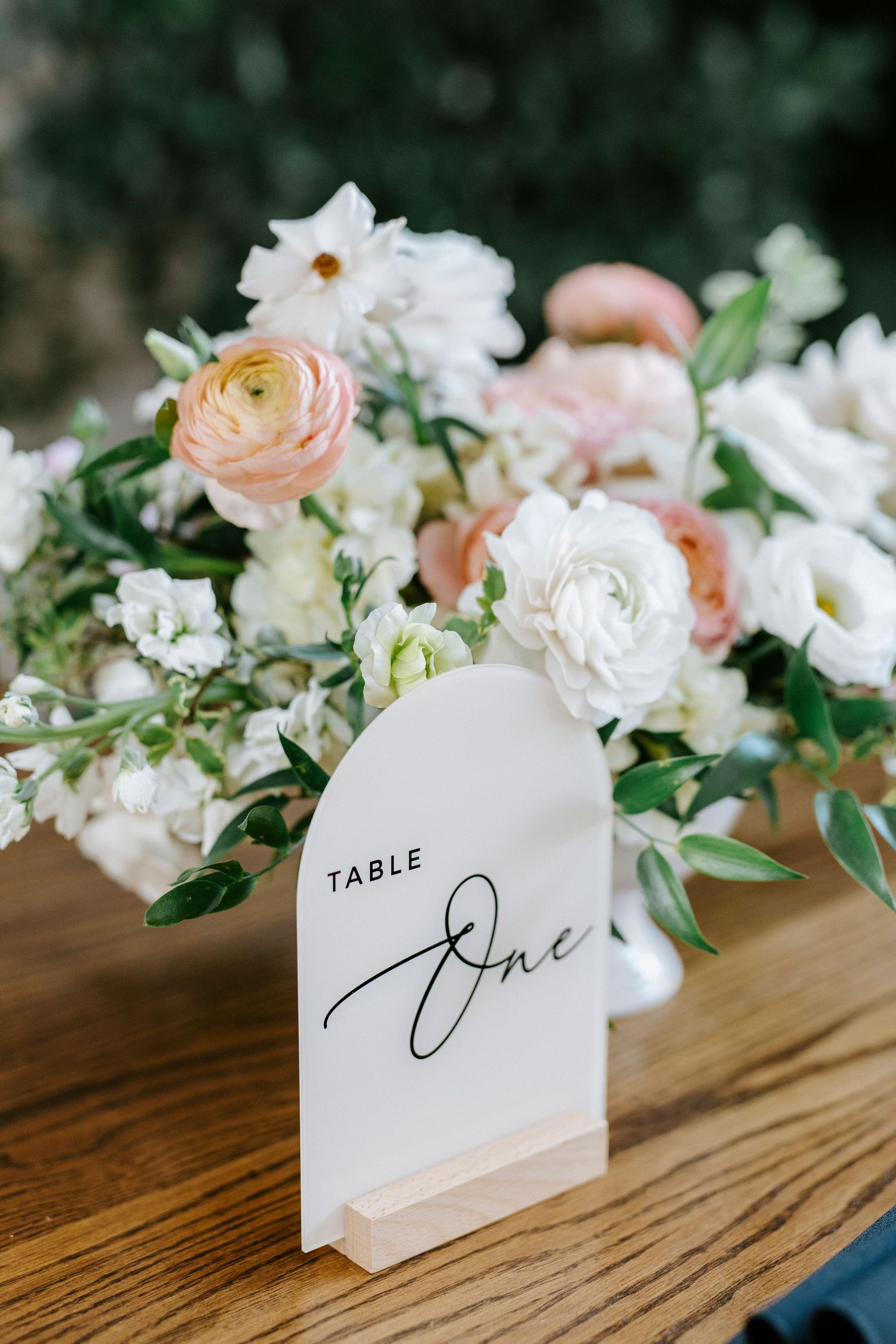 Elegant wedding table number card reading 'One' displayed among delicate white and blush floral arrangements on wooden surface.