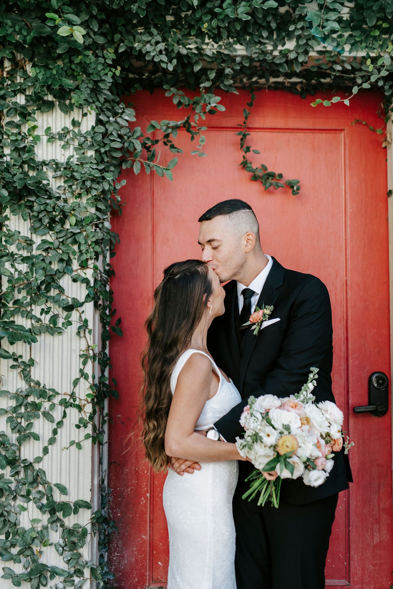 Newlyweds share romantic moments against a vibrant red door with climbing ivy.