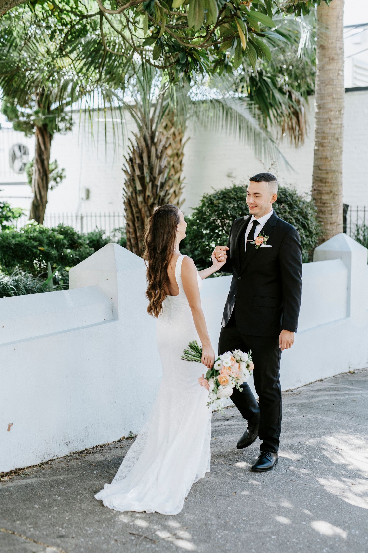 A wedding couple twirls and dances together on a palm-lined path with white walls and dappled sunlight.