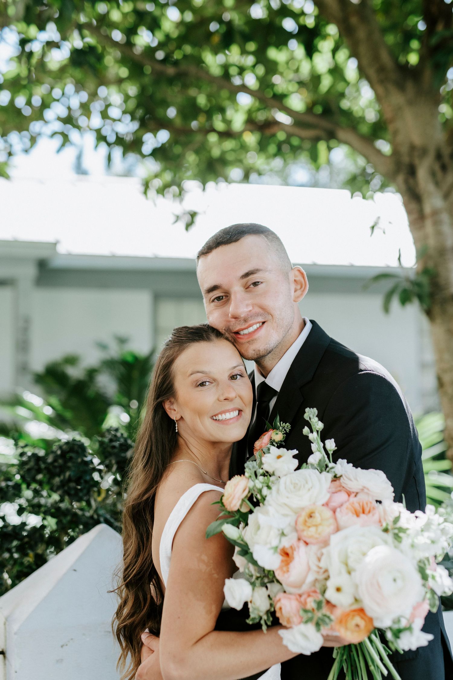 Newlyweds share a joyful moment outdoors while holding a beautiful bouquet of white and peach flowers.