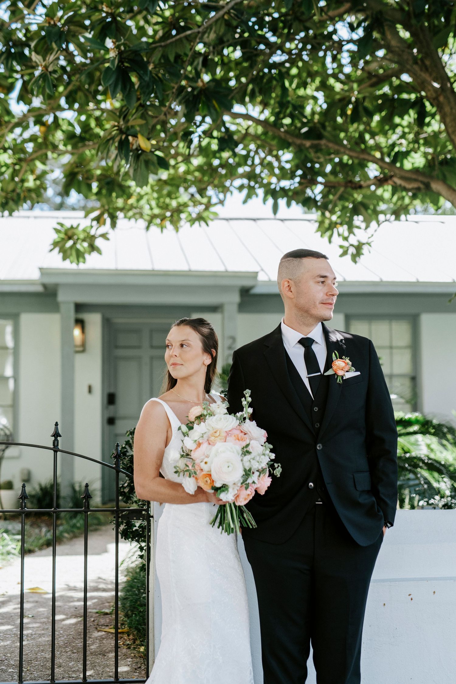 A bride and groom pose together outdoors under green trees, with the bride holding a pink and white floral bouquet.