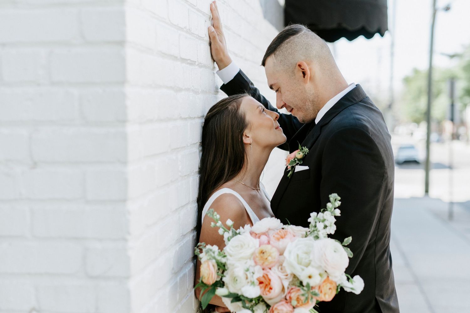Romantic outdoor wedding scene with couple embracing against a white wall background.