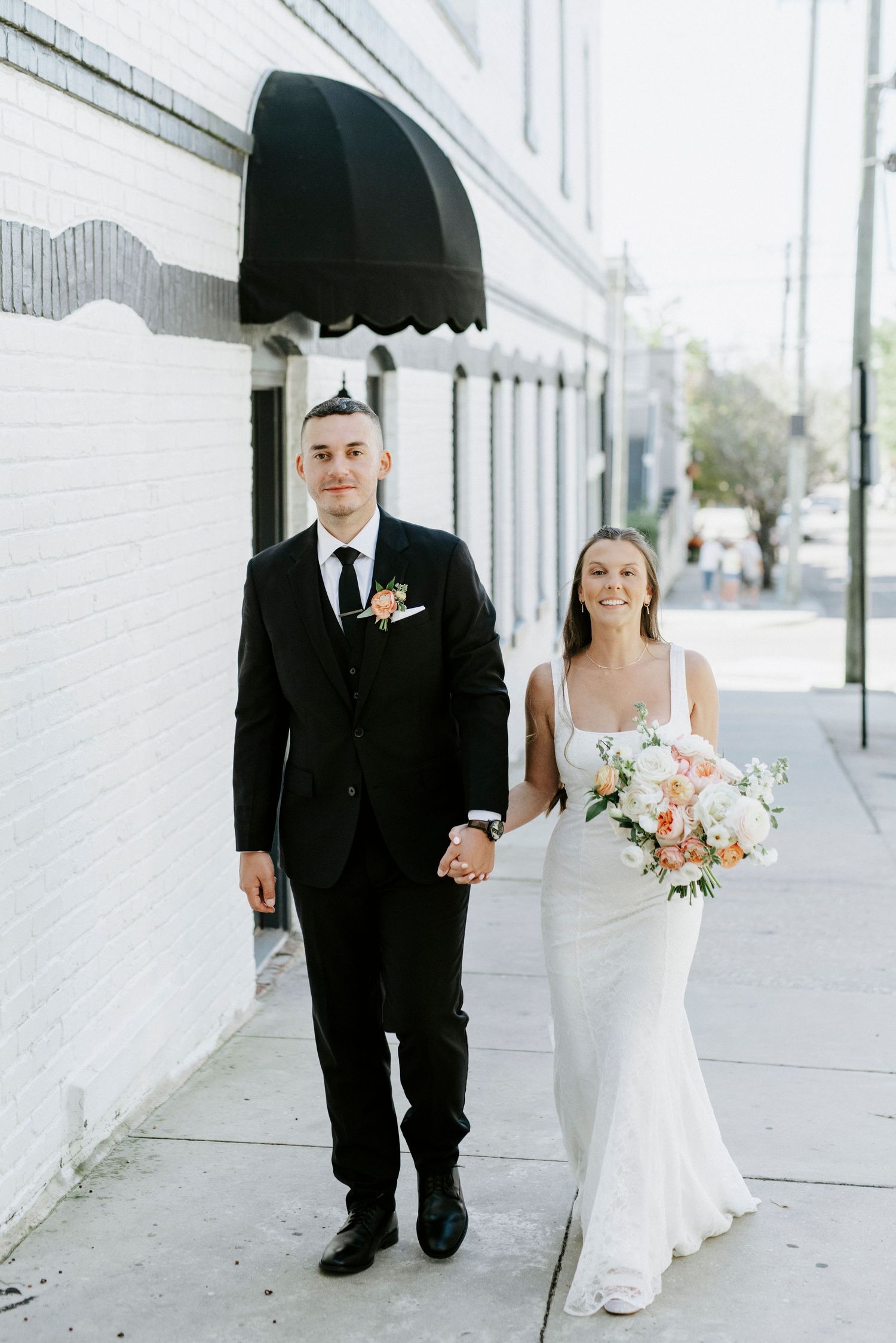 Wedding couple walks along white brick building exterior during their portrait session.