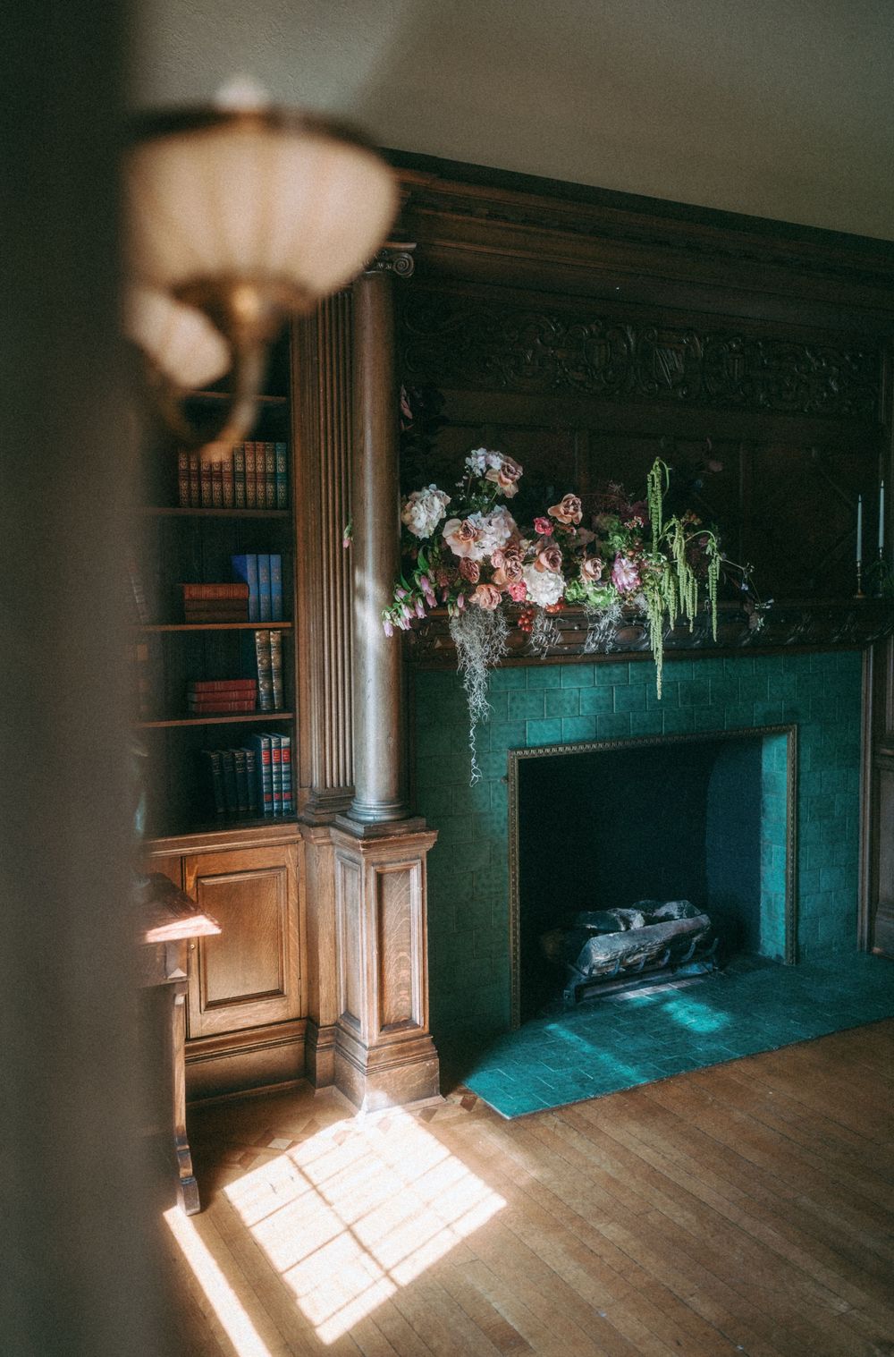 Sunlight streams through windows in a dark wooden paneled room with vintage furniture and hanging floral arrangements.
