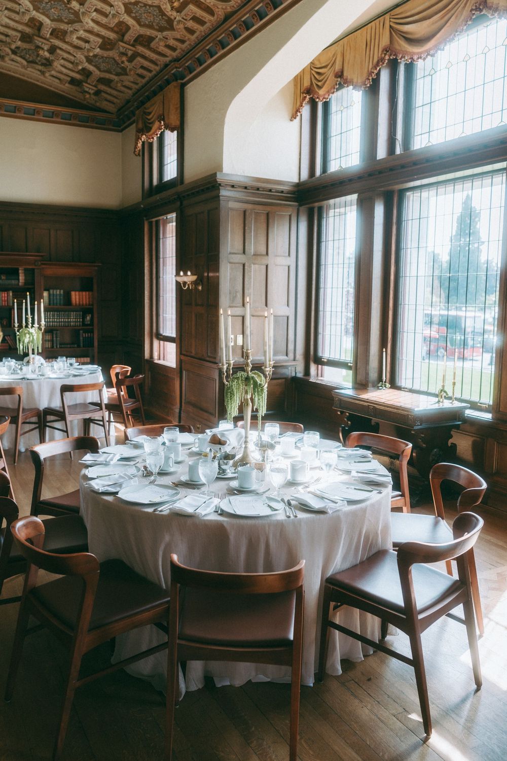 Elegant dining room setup with round tables covered in white linens and wooden chairs near large windows.