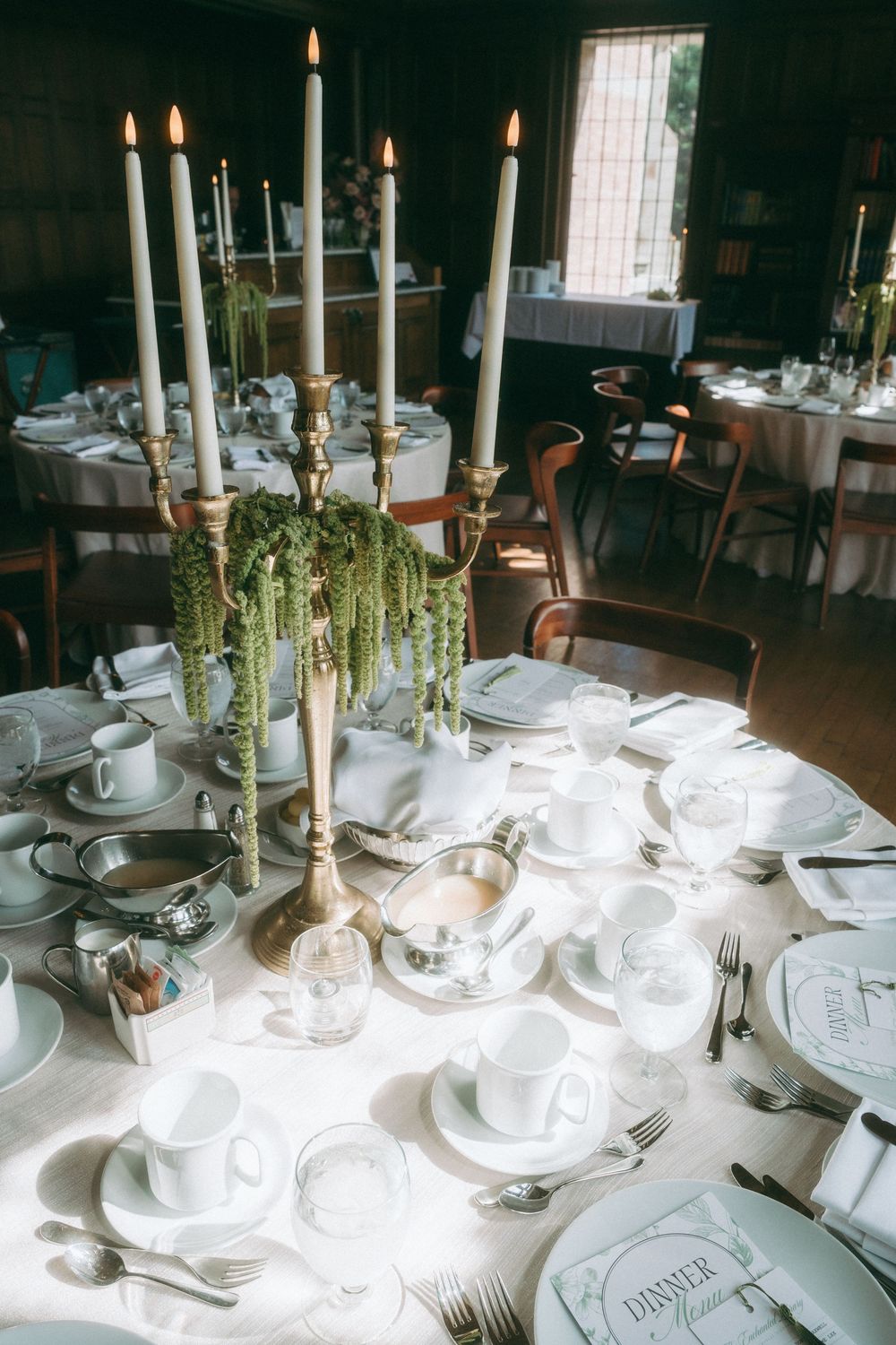 White place settings and tall candles arranged on formal dining tables in a dimly lit room.