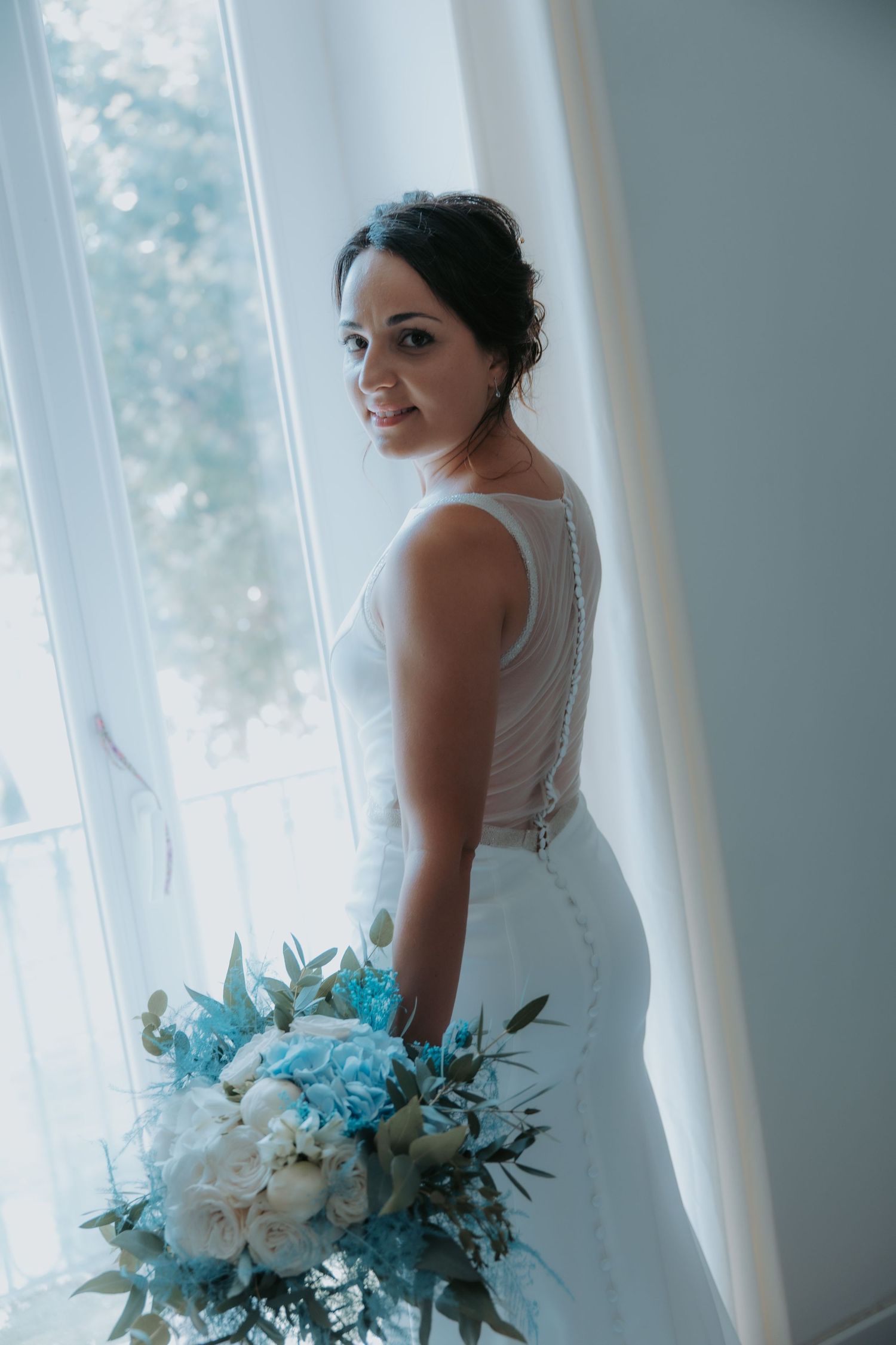 Elegant bride in white dress poses by window with blue and white floral bouquet.