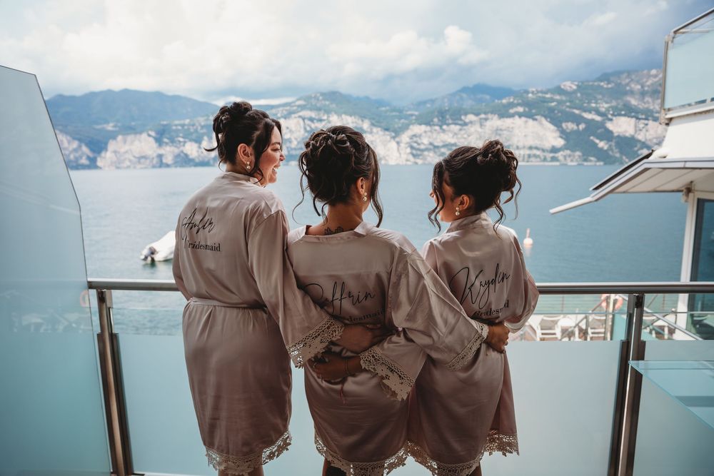 Bridesmaids in matching robes admire lake view from balcony.