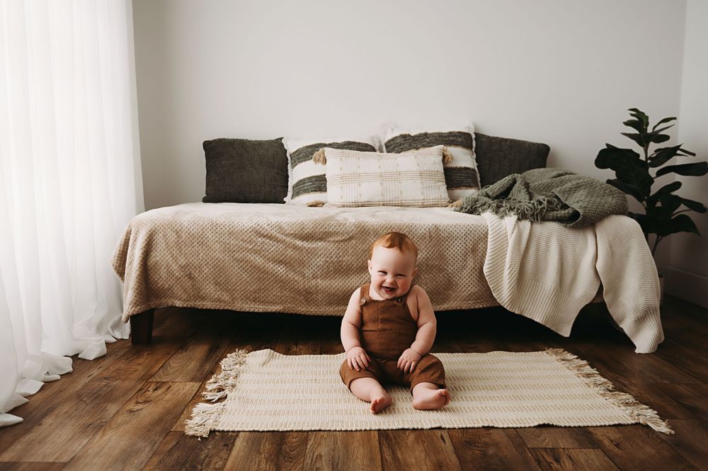 A small child sits on a woven rug in front of a cozy bed with neutral bedding and throw pillows.