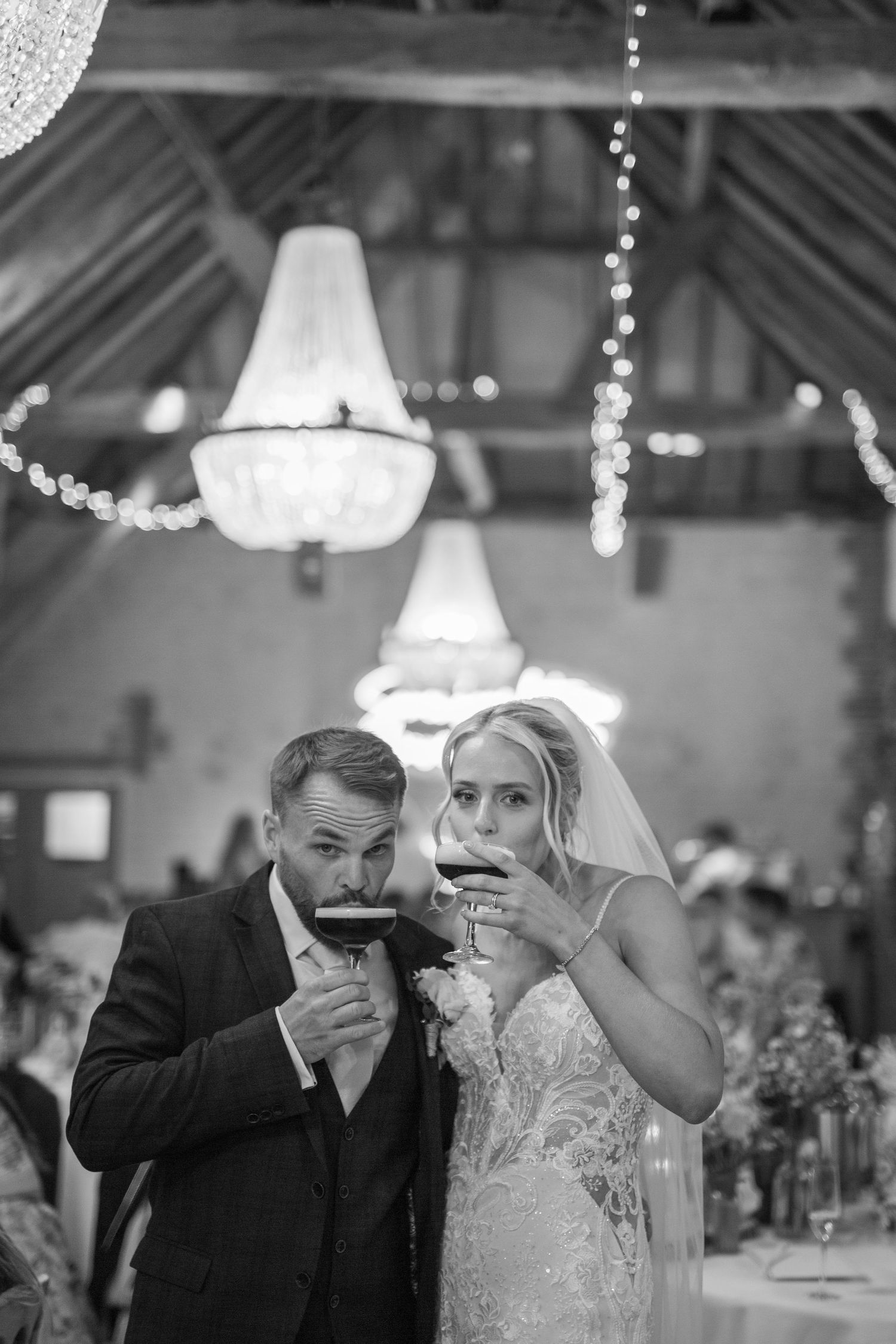 A couple in wedding attire sips drinks together under elegant chandeliers at an indoor reception venue.