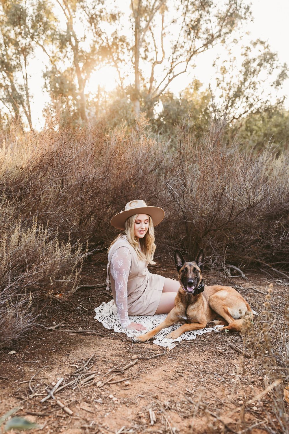 A Belgian Malinois dog rests on a blanket next to someone in a hat during golden hour in a rustic outdoor setting.