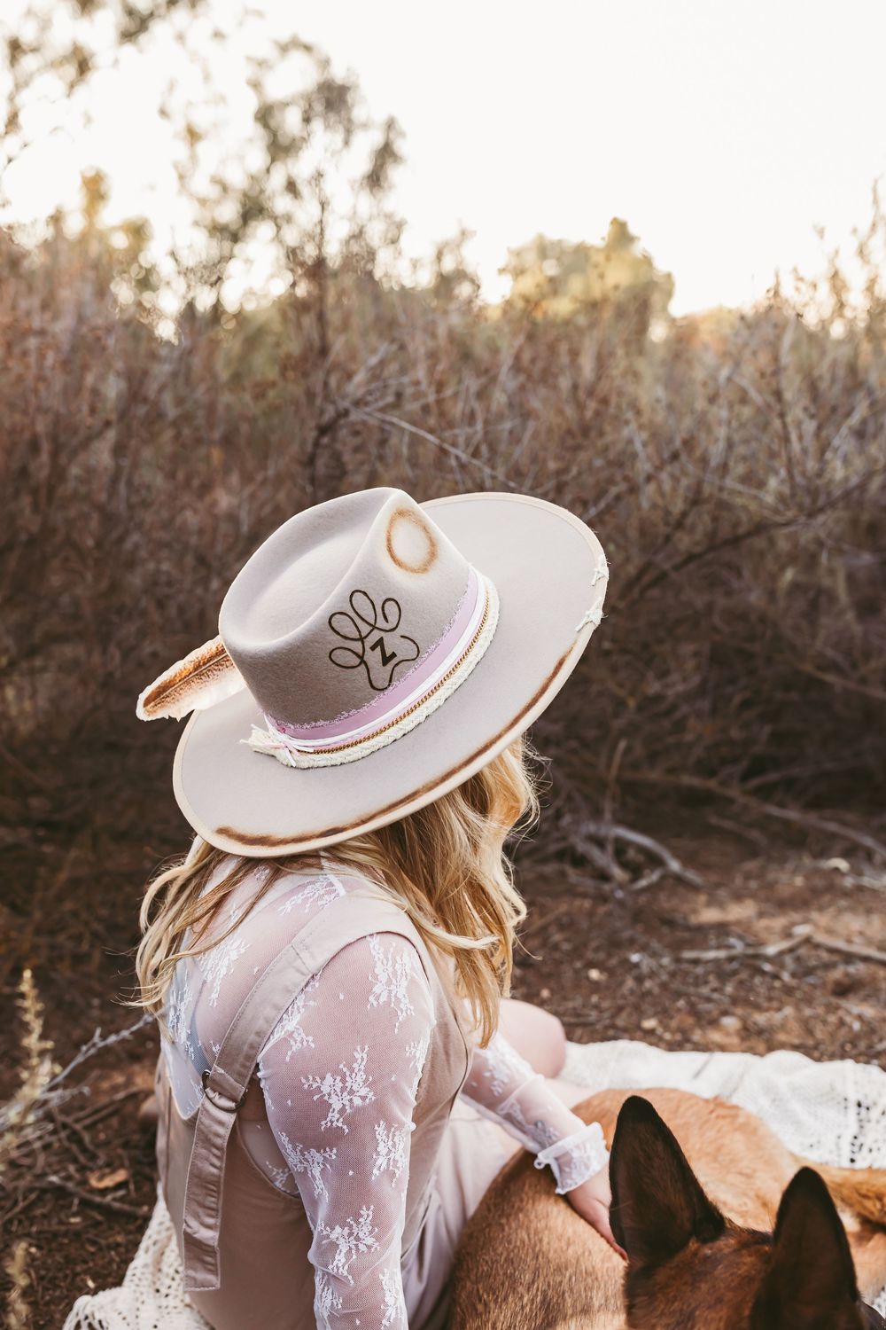 A person in a white lace dress and wide-brimmed hat sits outdoors during golden hour sunset.