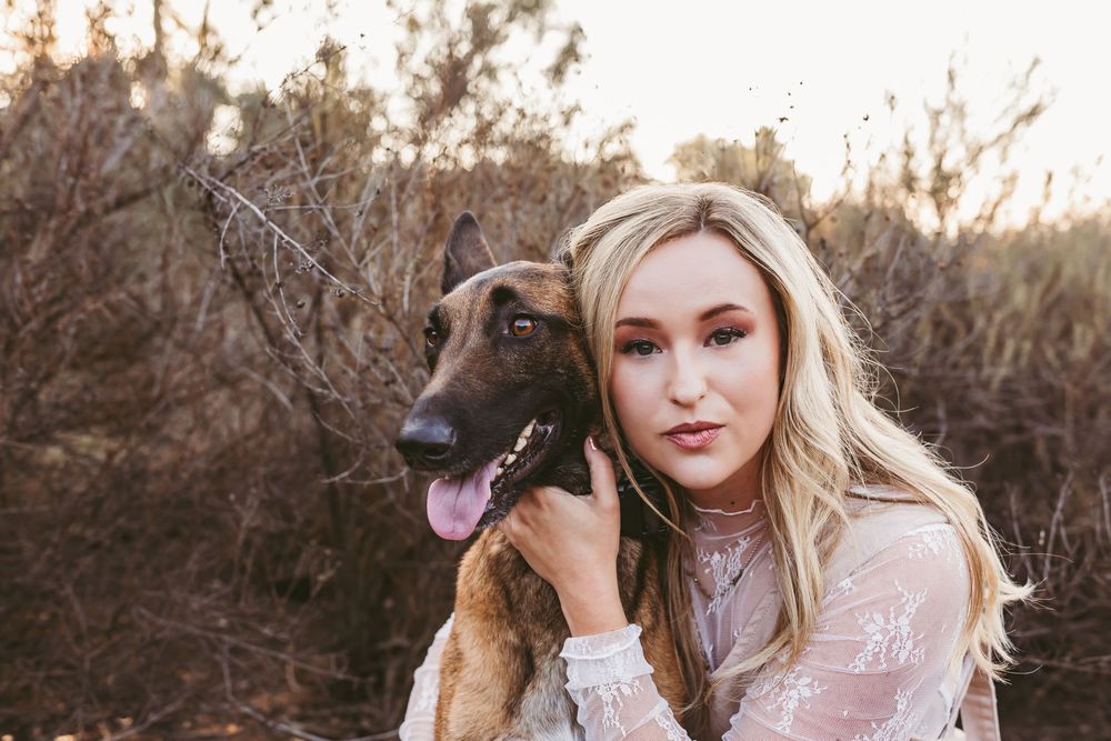 A Belgian Malinois dog poses outdoors with someone wearing a white lace dress during golden hour.