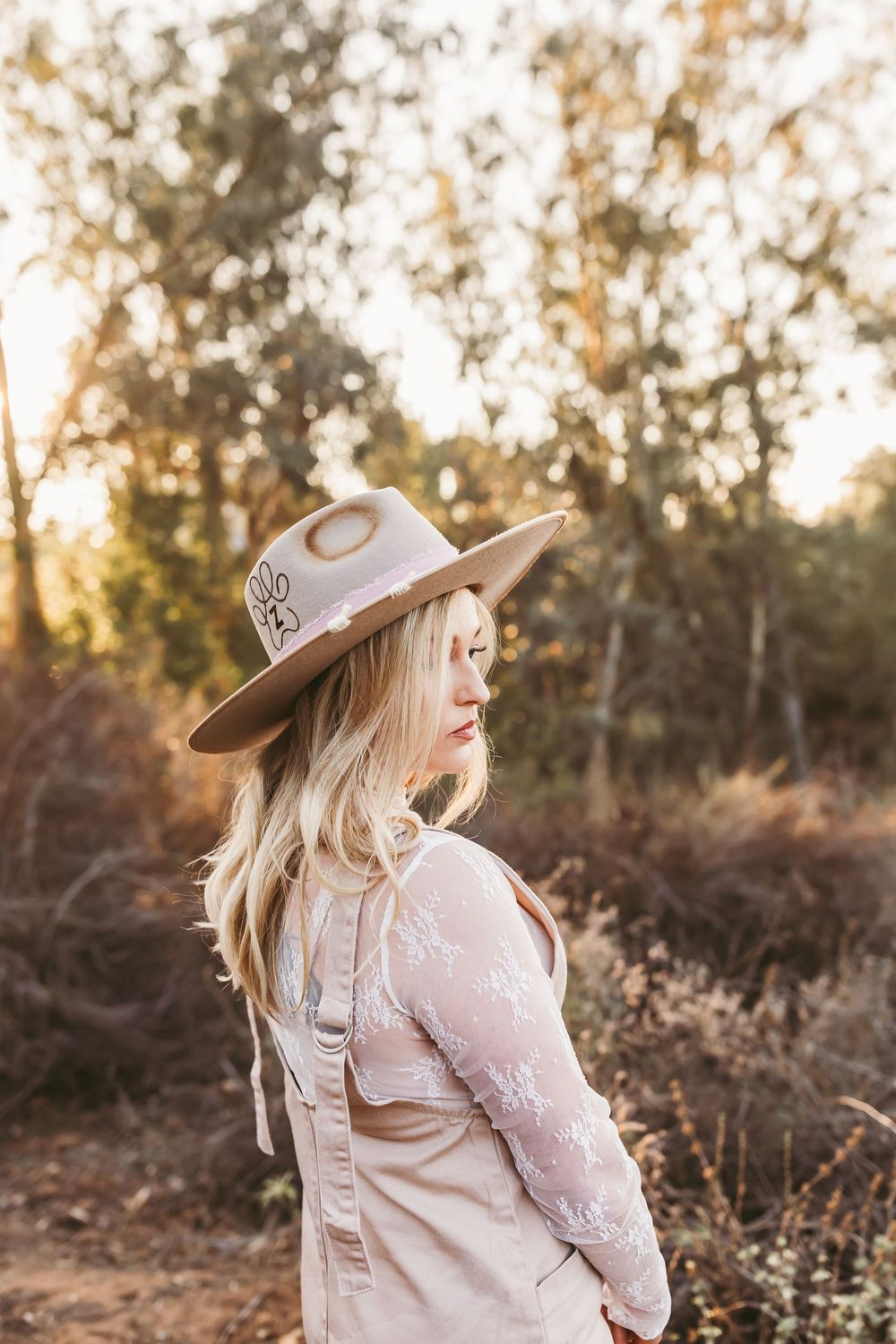 A person wearing a wide-brimmed hat and dress stands among brush during sunset.