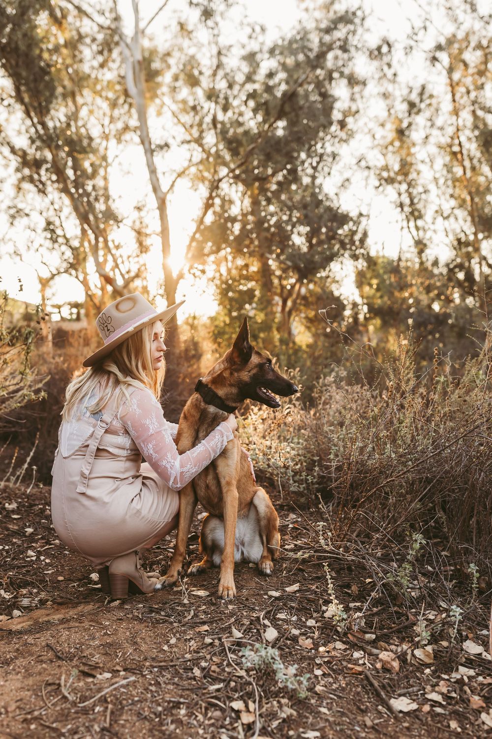 A person in a dress and hat kneels beside a German Shepherd dog in a sunlit natural setting.