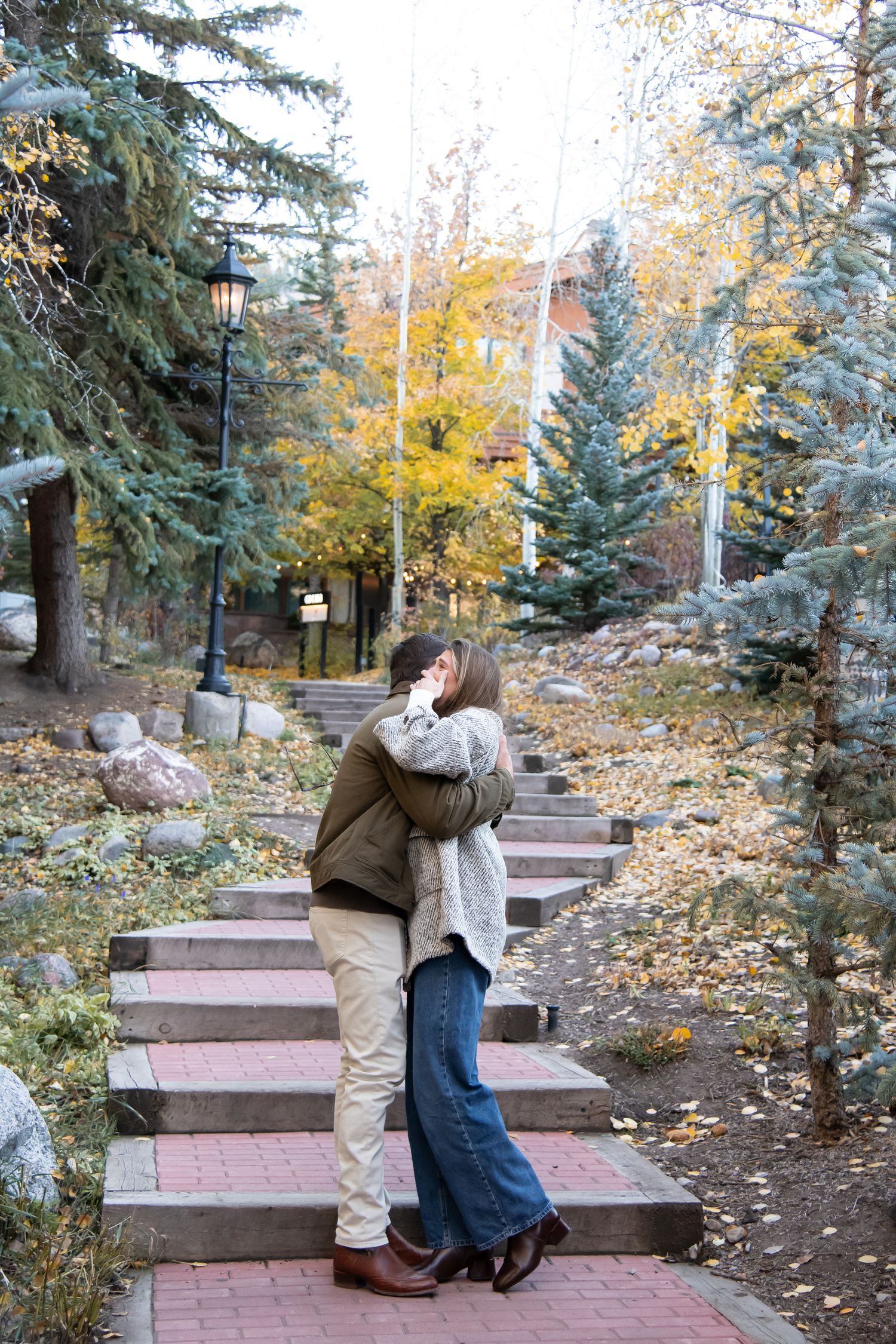 A cute couple embraces on romantic stairs with fall leaves in the background after a proposal in Vail Village Colorado
