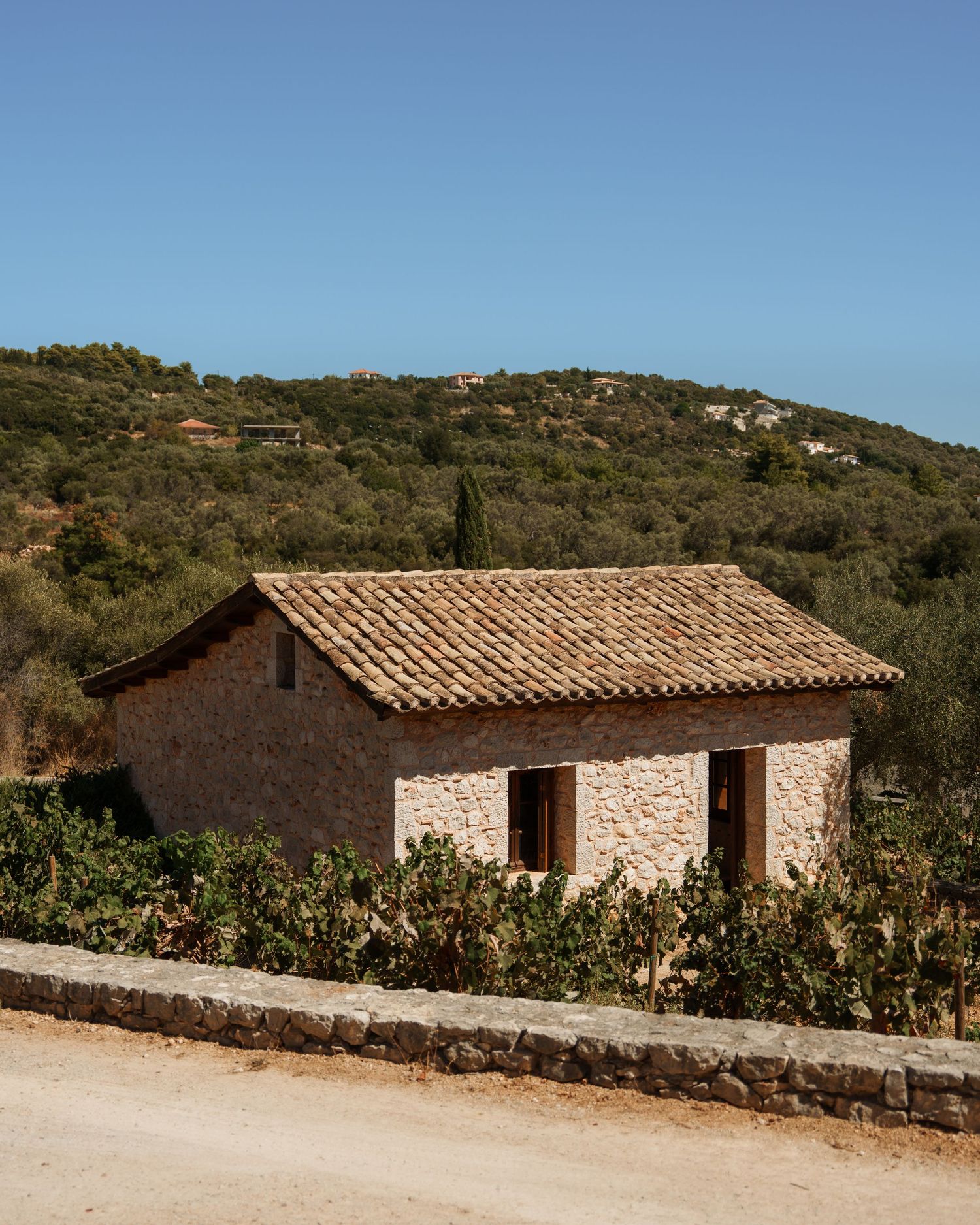 A row of rustic stone cottages with terracotta tiled roofs nestled against a hillside in a Mediterranean landscape.
