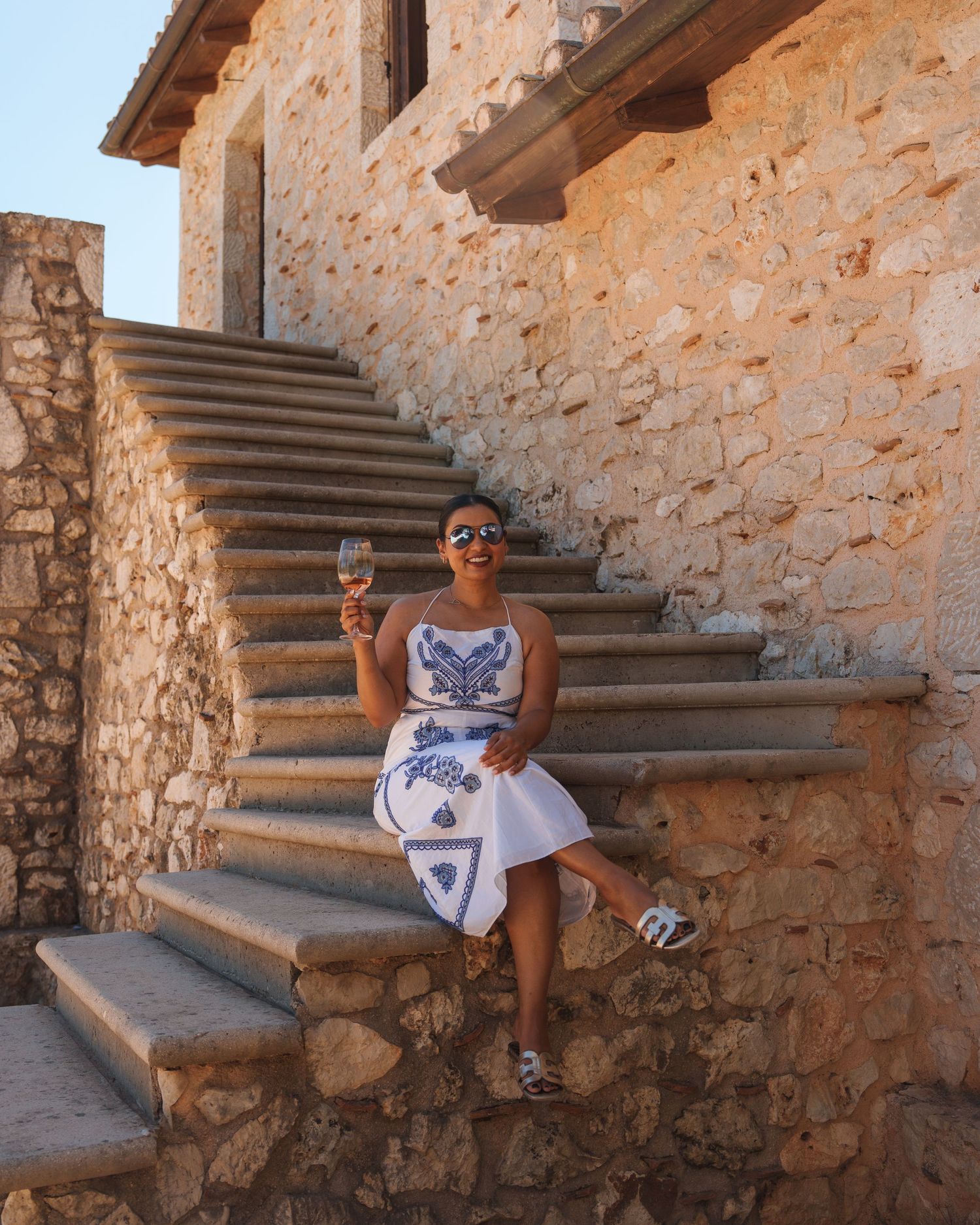 Someone sitting on stone steps against a rustic Mediterranean wall in a casual summer outfit.