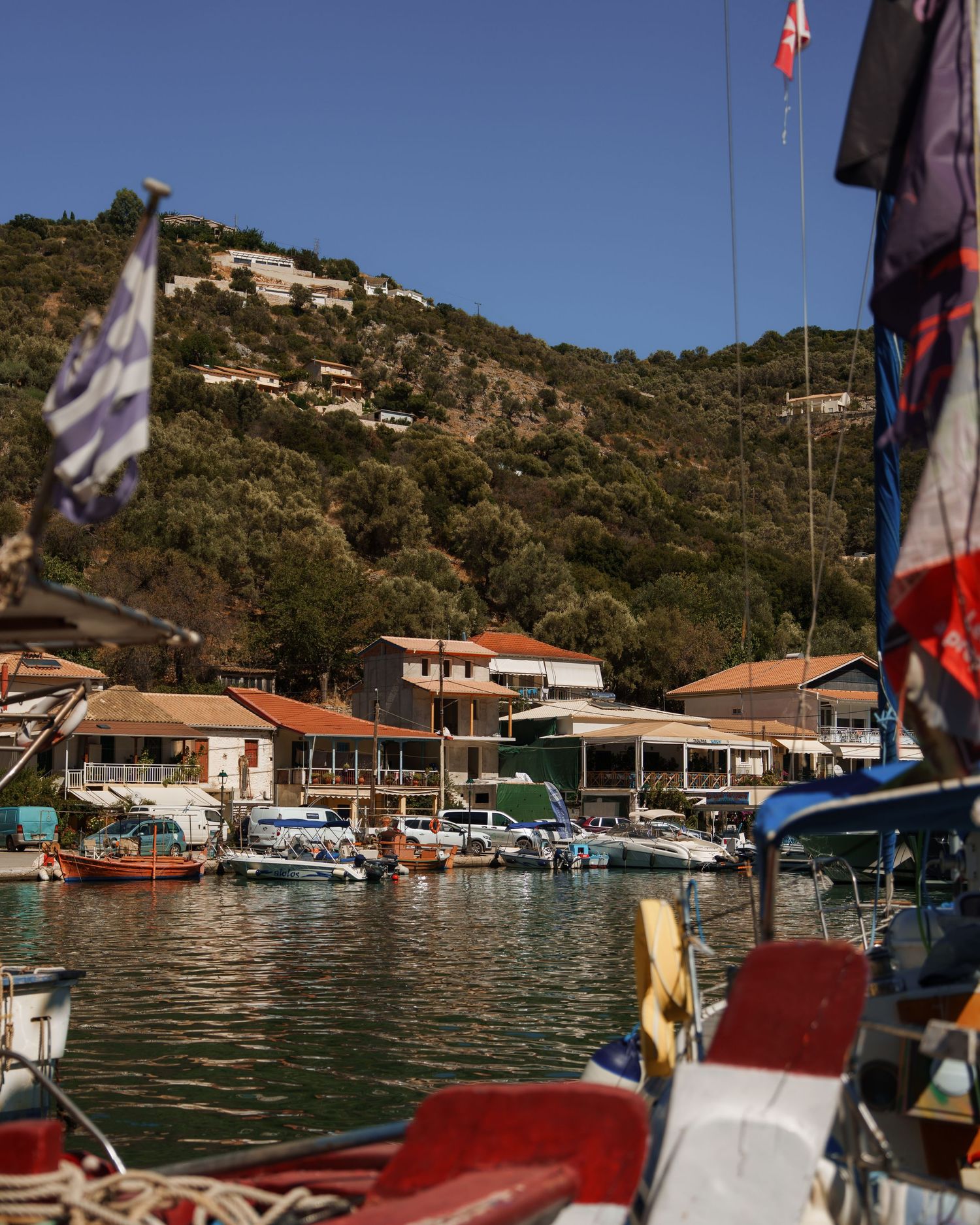 Small Mediterranean marina with boats docked at a waterfront village nestled against green hillsides under blue skies.
